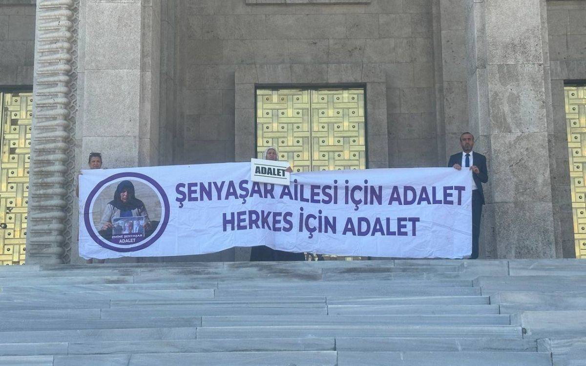 Bu fotoğrafta, Türkiye Büyük Millet Meclisi'nin önünde bulunan merdivenlerde iki kişi, ellerinde büyük bir pankart tutuyor. Pankartta, mor renkte büyük harflerle "Şenyaşar Ailesi İçin Adalet, Herkes İçin Adalet" yazısı yer alıyor. Pankartın sol tarafında, Emine Şenyaşar'ın bir fotoğrafı bulunuyor ve bu fotoğrafın altında "Emine Şenyaşar Adalet" ifadesi yazılı. Fotoğrafta, altın kaplamalı kapıların bulunduğu bir yapının önünde, kişilerden biri pankartın ortasında küçük bir "Adalet" yazılı levha tutuyor. Arka planda, bina ve merdivenlerin basamakları net bir şekilde görülüyor. Fotoğrafın genel atmosferi, adalet arayışını simgeliyor.