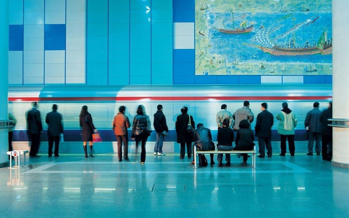 Bu fotoğrafta, bir metro istasyonunda bekleyen insanların görüntüsü yer alıyor. Ön planda, çeşitli yaş gruplarından insanlar ayakta duruyor veya bir bankta oturuyorlar. Ayakta duran kişiler genellikle sırtları kameraya dönük şekilde metro hattına bakıyorlar. Bankta oturanlar da metroyu izliyorlar. Arka planda mavi tonlarında döşenmiş bir duvar görülüyor, duvarın üzerinde ise eski deniz haritası ya da tarihi bir deniz sahnesini gösteren büyük bir tablo asılı.  Metro treni, fotoğraf çekilirken hareket halinde olduğu için biraz bulanık bir şekilde görünüyor, bu da hızlı hareket eden treni net bir şekilde gösteriyor. İstasyon oldukça temiz ve modern görünüyor, zemin parlıyor ve genel olarak düzenli bir ortam izlenimi veriyor. Ortamda soğuk tonlar hakim ve bu, ışıklandırma ve duvarların renginden kaynaklanıyor olabilir.  Fotoğrafta yer alan kişiler sakin ve bekleyiş içinde, günlük hayatın bir parçası olan metro yolculuğu için hazır durumda görünüyorlar