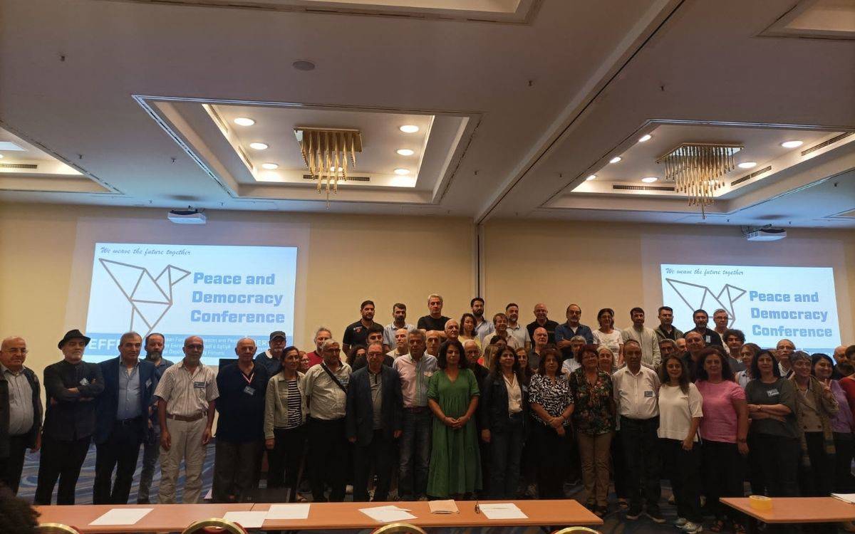 Bu fotoğrafta, geniş bir katılımcı grubu toplu halde poz veriyor. Grup, bir konferans veya seminer salonunda toplanmış gibi görünüyor. Arka planda, "Peace and Democracy Conference" (Barış ve Demokrasi Konferansı) yazılı bir projeksiyon ekranı bulunuyor. Bu, etkinliğin ana temasını ve amacını yansıtıyor.  Katılımcılar arasında kadınlar ve erkekler yer alıyor; çeşitli yaş gruplarından insanlar bir arada. Çoğu kişi gündelik kıyafetler giymiş, bazıları ise daha resmi giyinmiş. Ortam, dostane ve kolektif bir ruhu yansıtıyor.  Salonda sade bir dekorasyon mevcut; beyaz duvarlar, sade avizeler ve kırmızı sandalyeler dikkat çekiyor. Grup, fotoğraf çekimi için topluca kameraya bakıyor ve bazıları gülümseyerek poz veriyor. Bu fotoğraf, etkinliğe katılanların birlikte hareket ettiklerini ve barış ile demokrasi konularında ortak bir duruş sergilediklerini göstermekte.