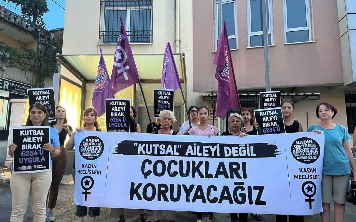 Bu fotoğrafta, bir grup kadın aktivist, bir protesto sırasında ellerinde pankartlar ve dövizler taşıyor. Pankartta büyük harflerle "Kutsal Aileyi Değil Çocukları Koruyacağız" yazıyor. Kadınlar, üzerinde "Kutsal Aileyi Bırak, 6284'ü Uygula" yazılı dövizler taşıyorlar. Arka planda ise birkaç bina ve bir kuaför dükkanı tabelası görülüyor. Aktivistlerin ellerinde Kadın Cinayetlerini Durduracağız Platformu ve Kadın Meclisleri logolarının bulunduğu bayraklar ve dövizler var. Fotoğraf, açık havada, bir sokakta çekilmiş ve eylemciler toplu halde durarak mesajlarını iletmeye çalışıyorlar.