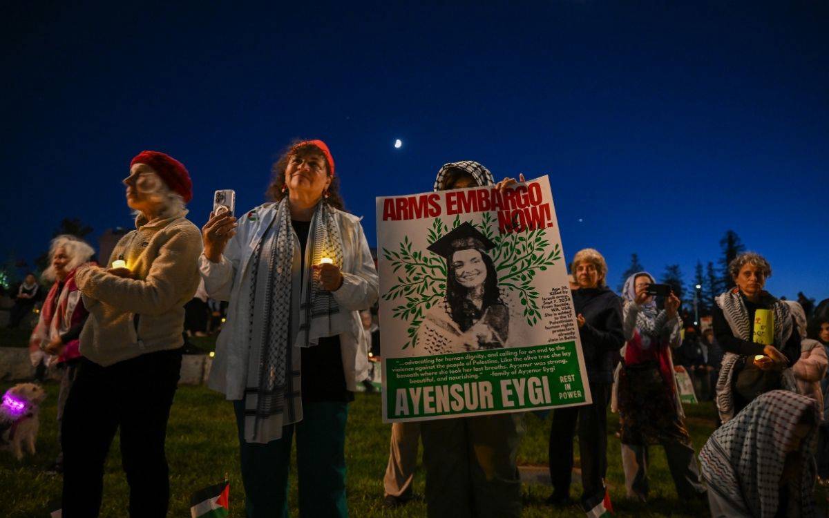 Bu fotoğrafta gece vakti, açık havada düzenlenen bir etkinlik ya da protesto görülüyor. Katılımcıların çoğu ellerinde mumlar taşıyor ve çeşitli pankartlar tutuyor. Fotoğrafın merkezinde bir kadın, üzerinde "ARMS EMBARGO NOW" (Silah Ambargosu Şimdi) yazan büyük bir pankart tutuyor. Pankartta ayrıca, mezuniyet kepi giymiş bir kadının fotoğrafı ve altında "Ayensur Eygi" ismi yer alıyor. Bu kişi muhtemelen etkinliğin odak noktası olan bir figür. Fotoğraftaki insanlar, silah ambargosu veya benzeri bir politik tepkiye yönelik farkındalık yaratmaya çalışıyor olabilir. Etkinliğe katılanlar genelde orta yaşlı veya yaşlı bireylerden oluşuyor ve ortam ciddi bir atmosfer taşıyor. Gökyüzünde ay ve yıldızlar görülebiliyor, bu da etkinliğin geç saatlerde düzenlendiğini gösteriyor.