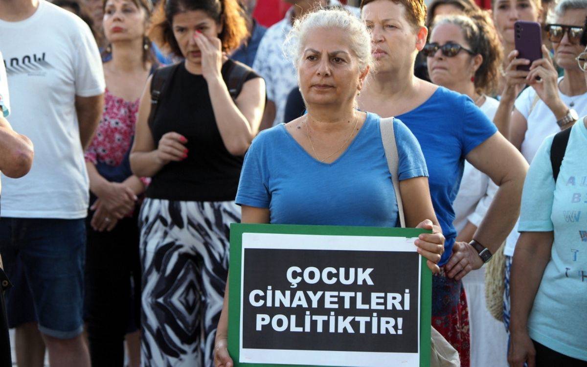 Bu fotoğrafta bir grup insan, bir protesto veya farkındalık etkinliğinde toplanmış durumda. Ön planda, mavi tişört giymiş beyaz saçlı bir kadın elinde bir pankart tutuyor. Pankartta, "Çocuk Cinayetleri Politiktir!" yazısı yer alıyor. Kadının yüz ifadesi ciddi ve kararlı görünüyor. Arka planda ise başka protestocular bulunuyor; bazıları telefonlarıyla fotoğraf çekiyor, bazıları düşünceli bir ifadeyle duruyor. Katılımcıların yüz ifadeleri, etkinliğin ciddiyetini ve önemini yansıtıyor. Topluluk, çocuk cinayetleri konusunda toplumsal bir mesaj vermeye çalışıyor olabilir.