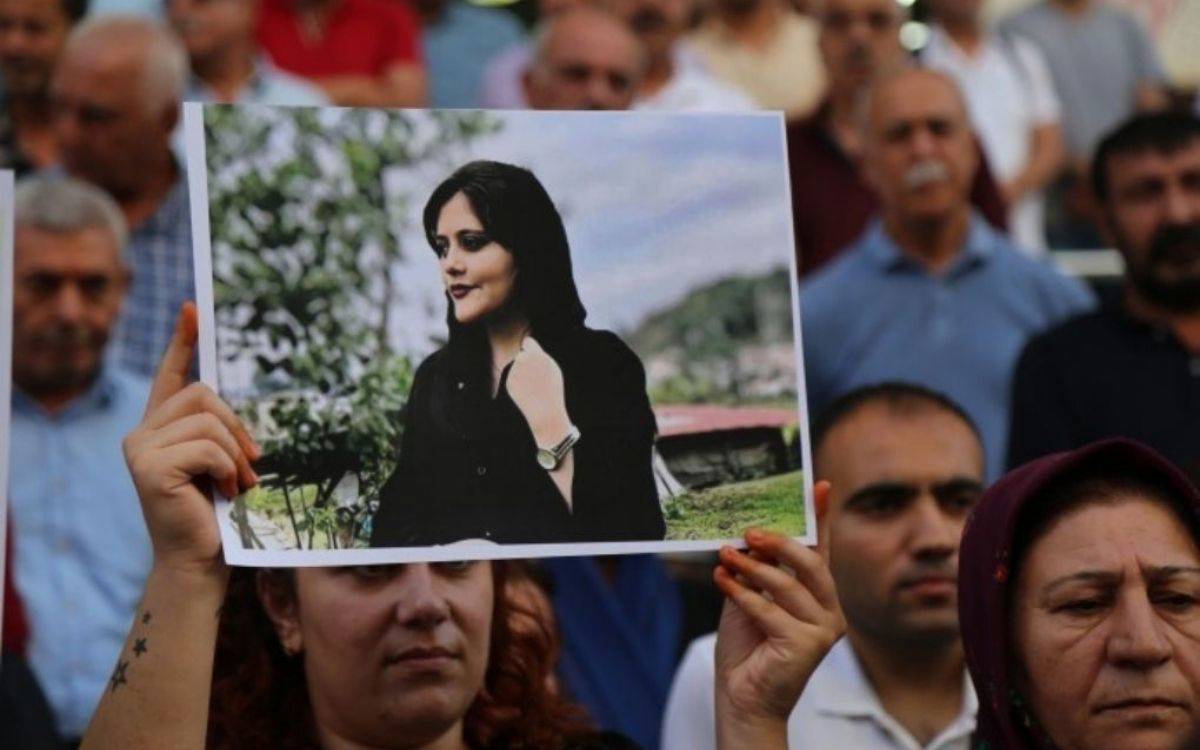 Bu fotoğrafta, bir grup insan açık bir alanda toplanmış durumda. Ön planda bir kadın, elleriyle Mahsa Amini'ye ait bir fotoğraf tutuyor. Fotoğrafın merkezindeki kadın, siyah bir giysi içinde, rahat ve doğal bir poz vermiş şekilde görülüyor. Arka planda ise bulanık bir şekilde duran, çoğunlukla orta yaşlı erkeklerden oluşan bir kalabalık mevcut. İnsanların yüz ifadeleri ciddiyet ve üzüntü yansıtıyor.