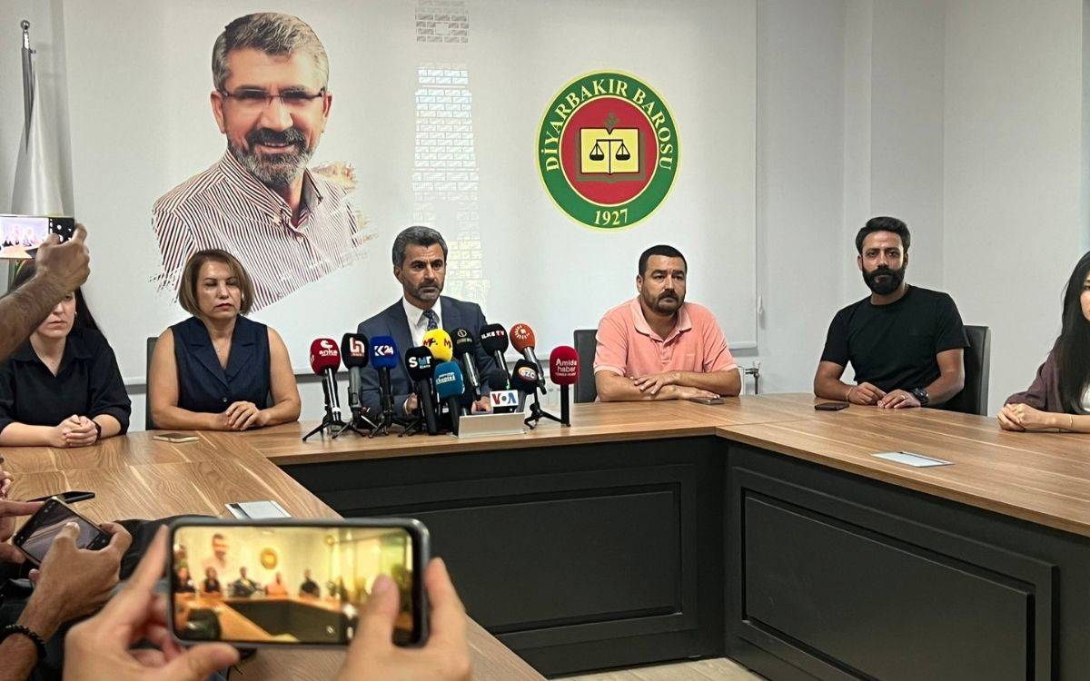 Fotoğrafta, Diyarbakır Barosu'nda gerçekleştirilen bir basın toplantısı yer alıyor. Masada oturan altı kişi dikkat çekiyor; merkezde takım elbiseli bir adam ve etrafında sivil kıyafetli kişiler bulunuyor. Masanın üzerinde çeşitli medya kuruluşlarına ait mikrofonlar yerleştirilmiş. Toplantıyı izleyen kişiler, ellerindeki telefonlarla görüntü alıyor.