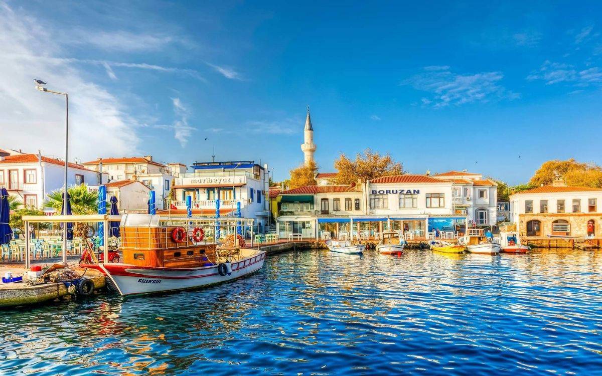 Bu fotoğraf, bir sahil kasabasının sakin ve huzurlu atmosferini yansıtmaktadır. Ön planda, suyun üzerinde birkaç küçük tekne yer alıyor. Özellikle ahşap gövdeli "Gülengül" adlı tekne dikkat çekiyor. Su berrak ve güneş ışığının yansımasıyla parlak bir mavi tonunda. Arka planda ise sahil boyunca dizilmiş beyaz ve pastel tonlarda binalar görülüyor. Binaların önünde restoran veya kafelere ait olduğu anlaşılan mavi-beyaz tenteler ve şemsiyeler var