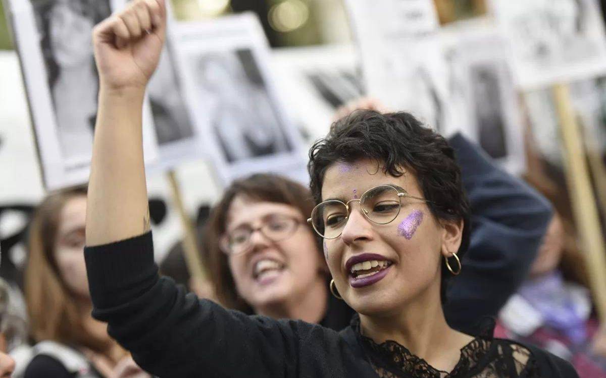 Bu görüntü, bir protesto veya toplumsal bir gösteriye katılan bir kişiyi gösteriyor. Odaktaki kişi, havaya yumruğunu kaldırmış, bu da genellikle dayanışma veya direnişin bir sembolü olarak kullanılır. Kişinin kısa, koyu renkli saçları, yuvarlak gözlükleri ve halka küpeleri var. Yüzünde mor renkli sim bulunuyor ve dudakları da mor rujla boyanmış, bu da ifade edici, kutlama ya da sembolik bir ruh hali yansıtır gibi görünüyor. Arkada başka kişiler de var ve ellerinde pankartlar ya da afişler tutuyorlar, ancak bu pankartların üzerindeki yazılar ya da görseller net bir şekilde seçilemiyor. Genel atmosfer, canlı ve dinamik, bir araya gelmiş toplu bir aktivist etkinliği izlenimi veriyor.