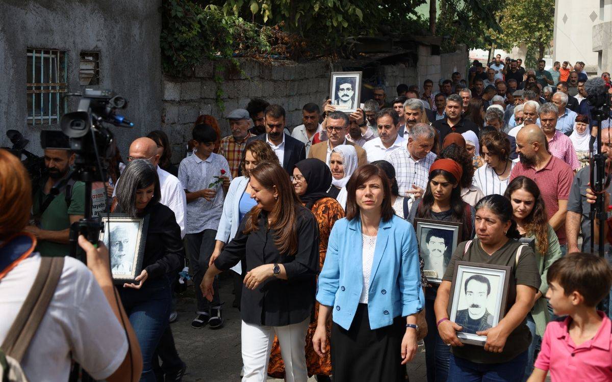 Bu fotoğraf, büyük bir grup insanın katıldığı bir yürüyüş veya anma törenini gösteriyor. Kalabalık arasında, önde yürüyen kişilerden bazıları ellerinde, çerçeveli siyah-beyaz portre fotoğraflar tutuyor. Bu, muhtemelen kaybedilen veya anılan kişilere atıfta bulunuyor. Fotoğraftaki kişiler, genellikle ciddi yüz ifadeleriyle dikkat çekiyor. Katılımcılardan bazıları ellerinde kırmızı karanfiller taşıyor.  Önde yer alan kadınlardan biri mavi bir ceket giymiş ve ciddi bir şekilde öne bakıyor. Yanında başka bir kadın, siyah giysiler içinde ve o da bir portre taşıyor. Geri planda farklı yaş gruplarından erkekler, kadınlar ve çocuklar da bu yürüyüşe katılmış. Ortamda ayrıca medya mensuplarının da bulunduğu görülüyor; kameralar ve fotoğraf makineleri dikkat çekiyor.  Yürüyüş, yoğun bir duygusal atmosferde, kayıp yakınlarını anma veya bir hak talebi etkinliği olabilir. Fotoğraf, bir dayanışma veya toplumsal adalet çağrısı içeren bir etkinlik gibi görünüyor.