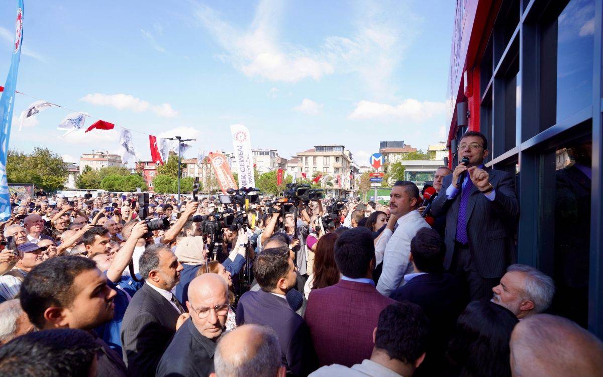 Bu fotoğrafta, büyük bir kalabalık dış mekânda toplanmış ve bir etkinlik sırasında konuşma yapan bir kişiyi dinliyor. Kalabalığın ortasında basın mensupları, kameralar ve fotoğraf makineleri ile konuşmacıyı takip ediyor. Konuşmayı yapan kişi, kalabalığın üzerinde yükseltilmiş bir platformda duruyor ve mikrofonla hitap ediyor. Konuşmacı, takım elbise giymiş ve etkinliği izleyen kalabalıkla iletişim kuruyor. Kalabalık oldukça yoğun ve dikkatle konuşmayı dinleyen insanlardan oluşuyor.  Arka planda çeşitli binalar, flamalar ve Türk bayrakları yer alıyor. Fotoğrafta ayrıca hava açık ve güneşli, gökyüzü mavi ve hafif bulutlu bir görünüme sahip. Alanda asılı olan flamalar ve kalabalığın heyecanlı duruşu, büyük bir miting veya bir açılış etkinliğini andırıyor.