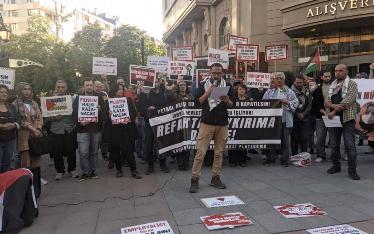 Fotoğrafta, açık alanda düzenlenen bir protesto veya basın açıklaması görülüyor. Ön planda bir kişi mikrofonla konuşma yaparken, arka planda ellerinde pankartlar tutan kalabalık bir grup yer alıyor. Pankartlarda "Filistin Halkı Kazanacak", "Emperyalist İsrail Kapatılsın", "Katliamcı İsrail İşbirlikçi AKP", "İsrail ile Ticaret Son Bulmalı" ve "Refah Tankeri Soykırıma Ortak" gibi Filistin’e destek ve İsrail’e karşı sloganlar dikkat çekiyor. Göstericiler, Filistin halkıyla dayanışma içinde olduklarını belirten çeşitli pankart ve dövizler taşıyor.