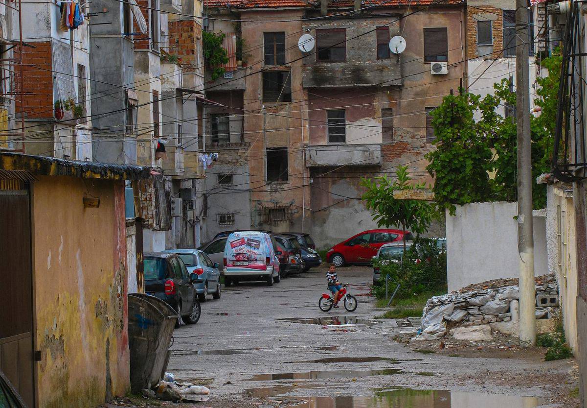 Görselde, eski ve bakımsız binaların bulunduğu bir mahalle görülüyor. Binaların cepheleri yıpranmış, sıvaları dökülmüş ve bazı yerlerde tuğlalar açıkta kalmış durumda. Apartmanların dış cephelerine asılmış çamaşırlar, balkonlarda uydu antenleri ve çok sayıda elektrik kablosu dikkat çekiyor. Sokakta çeşitli arabalar park etmiş ve bir çocuk kırmızı bir bisiklet sürüyor. Zemin oldukça düzensiz, yer yer su birikintileri ve çöpler mevcut. Bu görüntü, genel olarak yoksul bir mahalleyi ve bakımsız bir şehir manzarasını yansıtıyor.