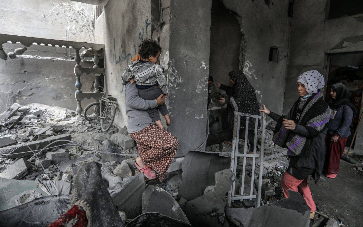 Fotoğrafta, bir kadın yıkılmış bir binanın içinde kucağında bir çocukla yürürken görülüyor. Kadın, gri bir üst ve desenli bir etek giymiş, kucağındaki çocuk ise gri bir kapüşonlu kıyafetle korunmuş durumda. Kadının yüzü görünmüyor, ancak dikkatle yürüdüğü belli oluyor. Çevrede büyük bir yıkım var; yerler molozlarla dolu ve duvarlarda ağır hasarlar mevcut. Arka planda iki kadın daha yıkıntılar arasında hareket ediyor, biri bir metal parçasını tutuyor. Yerde bisiklet, kırık parçalar ve yıkık bir balkonun parçaları bulunuyor. Fotoğraf, savaşın yıkıcı etkilerini ve sivil halkın bu zorlu koşullarda hayatta kalma mücadelesini çarpıcı bir şekilde yansıtıyor.