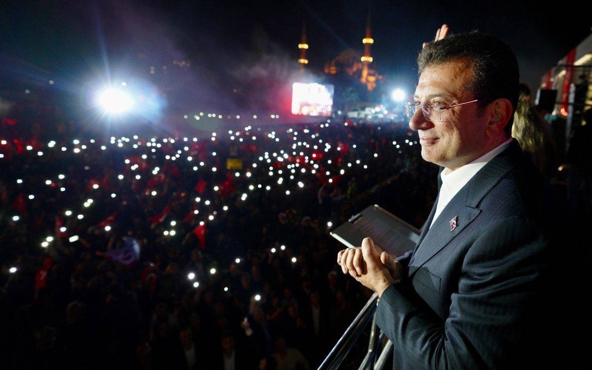 Bu fotoğrafta, İstanbul Büyükşehir Belediye Başkanı Ekrem İmamoğlu, gece saatlerinde bir kalabalığa hitap ederken görüntülenmiş. İmamoğlu, bir balkonda ya da yüksek bir platformda duruyor, elinde bir tablet veya not defteri tutuyor. Kalabalık, ellerindeki telefonların ışıklarıyla ortamı aydınlatmış durumda, coşkulu bir atmosfer hakim. Arka planda ise aydınlatılmış minareleriyle camiler görünüyor. Fotoğraf genel olarak miting havasını yansıtıyor ve İmamoğlu'nun kalabalık bir topluluğa hitap ettiği bir anı gösteriyor. Atmosferde hem bir politik konuşmanın ciddiyeti hem de destekleyici bir halk kitlesinin enerjisi hissediliyor.