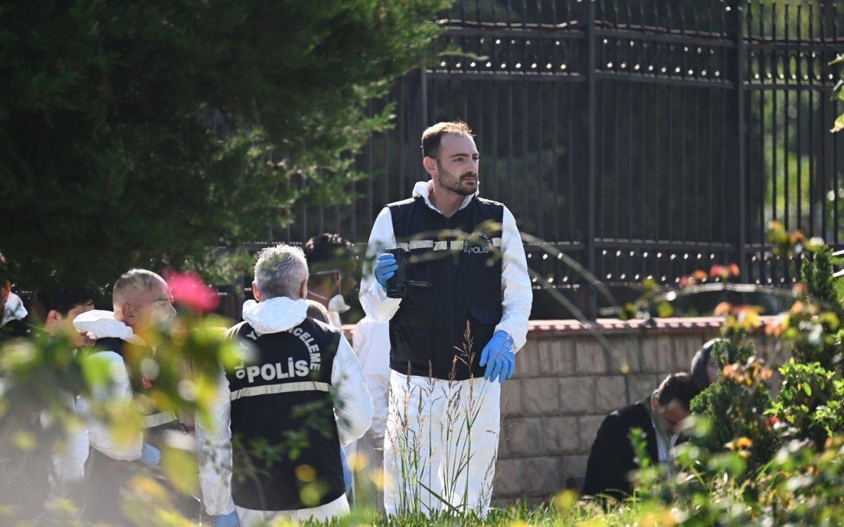 Fotoğrafta, olay yeri inceleme ekiplerinden bir grup polis, bir alanda inceleme yaparken görülüyor. Polis memurları, üzerlerinde "Olay Yeri İnceleme" yazılı yelekler ve beyaz koruyucu tulumlar giymişler. Önde bulunan polis memurlarından biri, elinde inceleme ekipmanları tutuyor ve mavi eldiven takmış durumda. Arka planda metal çitler ve yoğun ağaçlar bulunuyor, bu da olayın dış mekanda gerçekleştiğini gösteriyor.  Polisler yoğun bir şekilde alanı incelerken, dikkatlice delil topladıkları anlaşılıyor. Fotoğraf, ciddi bir olay sonrası olay yerinin detaylıca araştırıldığını ve kriminal ekiplerin profesyonel bir şekilde çalıştığını yansıtıyor. Ön planda bitki örtüsü ve çiçekler de dikkat çekiyor, bu da olayın yeşil bir alanda gerçekleştiğini düşündürüyor.