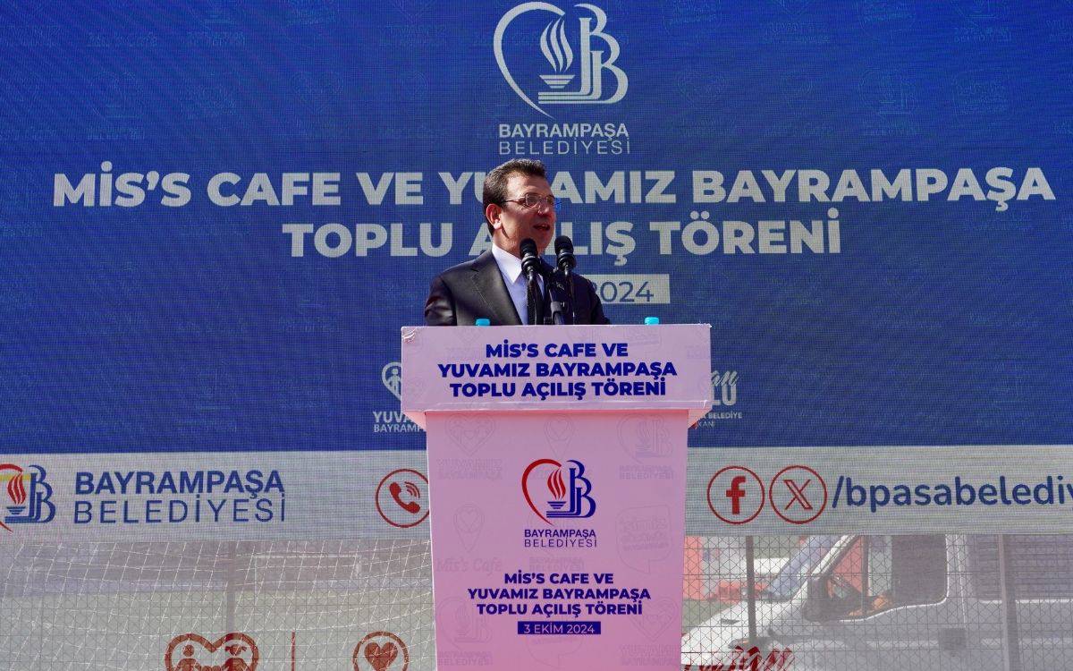 Fotoğrafta, İstanbul Büyükşehir Belediye Başkanı Ekrem İmamoğlu, Bayrampaşa Belediyesi’nin "Mis's Cafe ve Yuvamız Bayrampaşa Toplu Açılış Töreni"nde konuşma yaparken görülüyor. İmamoğlu, üzerinde etkinliğin adı yazılı bir kürsüde duruyor. Arka planda büyük bir afişte "Bayrampaşa Belediyesi" logosu ve etkinliğin ismi net bir şekilde yer alıyor. Kürsüde ise yine etkinliğin adı ve Bayrampaşa Belediyesi'nin logosu bulunmakta. İmamoğlu'nun konuşma yaptığı sırada resmi bir ortamda olduğu, takım elbise giymesi ve törendeki mikrofonlar dikkat çekiyor