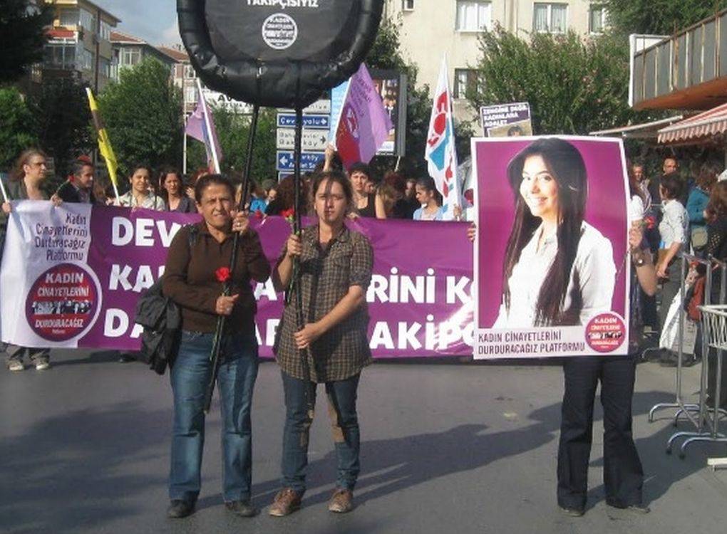 Fotoğrafta, "Kadın Cinayetlerini Durduracağız Platformu" tarafından düzenlenen bir protesto yürüyüşü görülüyor. Önde iki kişi yürümekte; biri elinde kırmızı bir karanfil tutuyor, diğeri ise büyük bir pankart taşıyor. Sağ tarafta, büyük bir pankart üzerinde bir kadının fotoğrafı yer alıyor ve bu pankartta "Kadın Cinayetlerini Durduracağız Platformu" ifadesi bulunuyor. Fotoğraftaki kadın, platformun destek verdiği bir cinayet kurbanına ait olabilir.