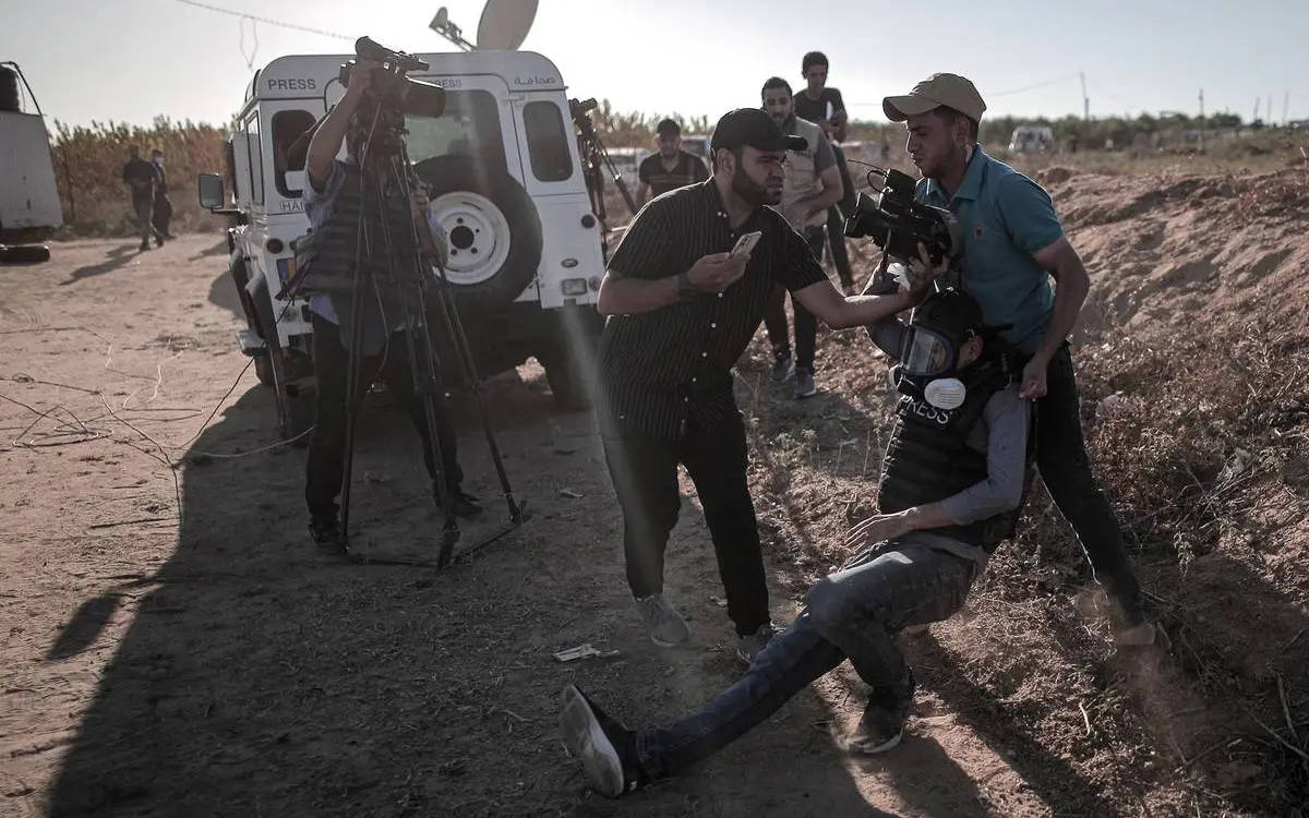 Fotoğrafta bir açık hava sahnesi görülüyor. Görüntüde birkaç kişi yer alıyor ve bu kişiler muhtemelen gazeteciler veya medya çalışanları. Ön planda, yere oturmuş olan bir kişi var; üzerinde "PRESS" (basın) yazılı bir yelek ve miğfer bulunuyor. Bu kişi muhtemelen yaralanmış, çünkü iki kişi onu kaldırmaya veya destek olmaya çalışıyor. Bir kişi miğferine tutunarak destek sağlıyor, diğer kişi ise yanından telefonla yardım çağırıyor gibi görünüyor.  Arka planda beyaz bir araç var, üzerinde de "PRESS" yazısı göze çarpıyor. Araç, bir medya aracı gibi görünüyor ve yanında kamera ekipmanları olan başka kişiler de var. Fotoğraftaki ortam toprak bir arazi, çalılar ve yol kenarına benzer bir yerleşimle çevrili. Işık, gün batımına yakın bir vakitte çekilmiş olduğunu düşündürüyor, çünkü gölgeler uzun ve ışık açısı düşük. Çevredeki hava gergin, acil bir durum yaşanıyor gibi görünüyor.