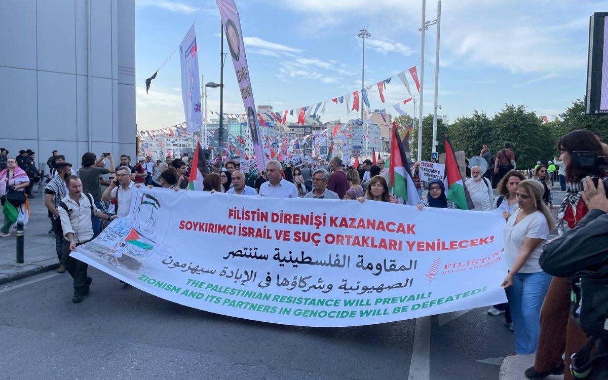 Fotoğraf, kalabalık bir grubun ellerinde pankartlarla bir yürüyüş gerçekleştirdiği bir anı göstermektedir. Yürüyüşe katılanlar, Filistin'e destek amacıyla toplandıkları görülmektedir. Önde büyük bir pankart taşınmakta olup, üzerinde üç dilde (Türkçe, Arapça ve İngilizce) mesajlar yer almaktadır. Türkçe kısımda "Filistin Direnişi Kazanacak, Soykırımcı İsrail ve Suç Ortakları Yenilecek!" yazısı öne çıkmaktadır. Pankartın İngilizce kısmında ise "The Palestinian Resistance Will Prevail! Zionism and Its Partners in Genocide Will Be Defeated!" ifadesi yer almaktadır.  Katılımcıların çoğunun Filistin bayrakları taşıdığı ve arka planda daha fazla kişinin olduğu görülmektedir. Yürüyüş sırasında ellerinde farklı pankartlar taşıyan insanlar da dikkat çekmektedir. Mekanın açık bir alanda, büyük bir meydanda olduğu anlaşılmaktadır ve gökyüzü açık ve güneşli bir hava hakimdir. Pankartların üzerindeki sloganlar, Filistin direnişine destek ve İsrail'e karşı tepki mesajları içermektedir.