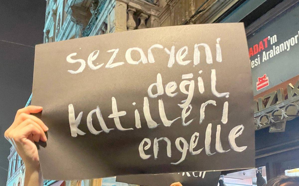 Görselde bir kişinin elinde tuttuğu siyah bir karton üzerinde, beyaz boya ile yazılmış bir mesaj yer alıyor. Kartonda "Sezaryeni değil katilleri engelle" yazıyor. Arka planda tarihi bir bina cephesi görülmekte, gece saatlerinde olduğu izlenimini veren bir atmosfer mevcut. Kartonun metni, kadın hakları veya toplumsal bir soruna dikkat çeken bir protesto niteliği taşıyor.