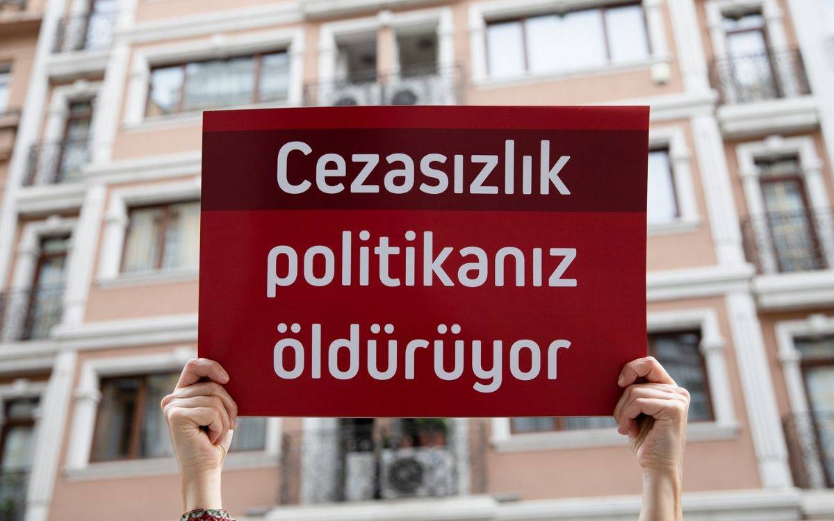 Bu fotoğrafta, bir kişinin elleriyle yukarıda tuttuğu bir pankart görülüyor. Pankartta, beyaz renkle yazılmış büyük harflerle "Cezasızlık politikanız öldürüyor" yazısı dikkat çekiyor. Arka planda, bir apartman binasının dış cephesi ve pencereleri görülüyor. Fotoğraf, bir protesto ya da eylemde çekilmiş olabilir, çünkü pankarttaki mesaj adalet sistemine yönelik bir eleştiriyi ifade ediyor. Yazı, toplumsal adalet ve cezasızlık politikasının kadın cinayetleri gibi şiddet olayları üzerindeki etkisine işaret ediyor.