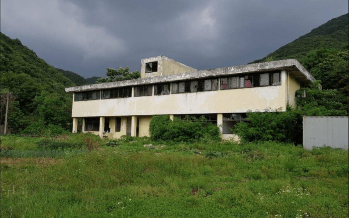 Fotoğrafta, doğa tarafından neredeyse sarılmış, terkedilmiş bir bina görülüyor. İki katlı, sararmış ve yıpranmış bir yapı. Üst kattaki pencereler hasarlı, camlar kırık ve yer yer demir parmaklıklar görünüyor. Duvarlar, dış koşulların etkisiyle yer yer soyulmuş ve dökülmüş. Çatı kısmında betonun aşındığı belli oluyor. Bina, yoğun yeşilliklerle çevrili bir alanda, arka planda ise dağlık bir bölge ve koyu bulutlarla kaplı bir gökyüzü dikkat çekiyor. Genel hava karamsar ve izole bir manzara sunuyor.