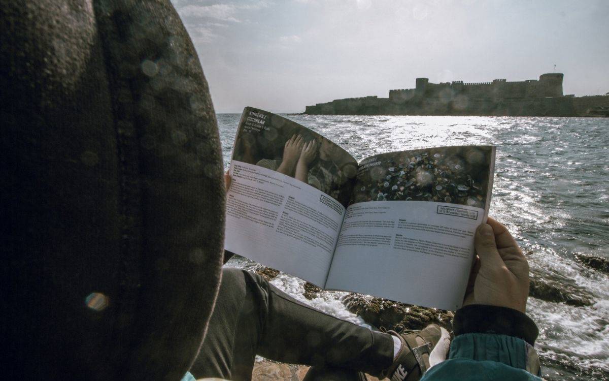 Görselde, bir kişi deniz kenarında oturmuş ve bir dergi ya da kitap okuyor. Kişi, kameraya sırtı dönük olarak görülüyor ve kucağındaki açık dergideki sayfaları inceliyor. Derginin sayfalarında metinler ve görseller yer alıyor. Derginin içeriği, elleri açık bir şekilde gökyüzüne uzanmış insanları gösteren bir görselle dikkat çekiyor. Kişinin başında bir kapüşon ya da bere var ve rahat bir şekilde deniz manzarasının keyfini çıkarıyor gibi görünüyor.