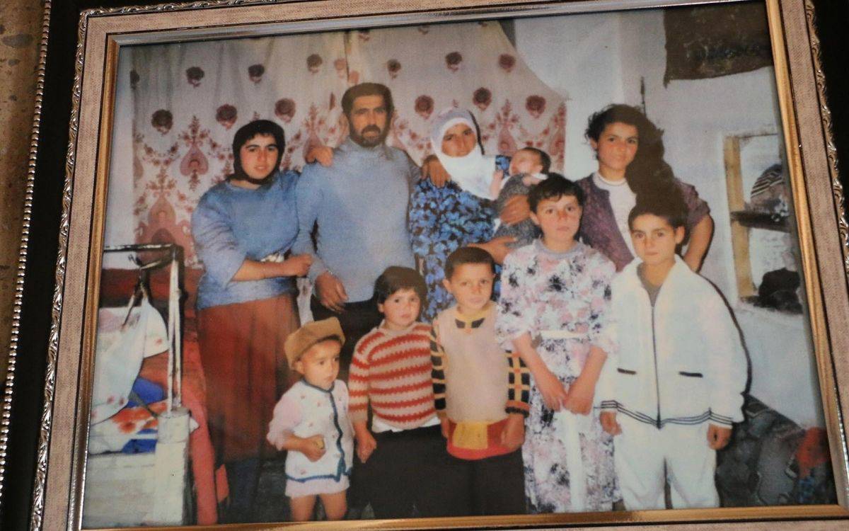 Bu fotoğrafta, bir aile iç mekanda birlikte poz veriyor. Aile üyeleri, geleneksel kıyafetler giymiş ve toplu halde, oldukça samimi bir ortamda duruyorlar. Arkada ahşap raflar üzerinde battaniyeler ve ev eşyaları yer alıyor, bu da ortamın bir ev içi yaşam alanı olduğunu gösteriyor. Ailede büyükler ve çocuklar birlikte, rahat bir pozda duruyorlar; bazı çocuklar elleriyle barış işareti yapıyor. Aile üyeleri birbirine yakın durarak sıcak ve koruyucu bir aile atmosferi yansıtıyor. Odadaki genel hava, sade ve mütevazı bir yaşam tarzını simgeliyor.