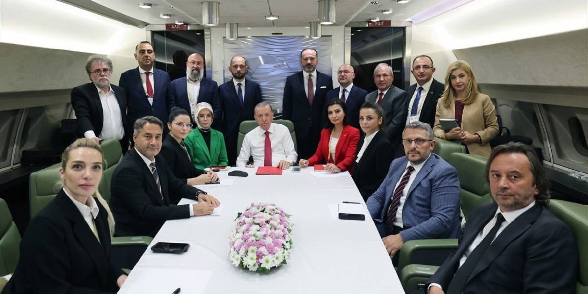 Bu fotoğraf, Cumhurbaşkanı Recep Tayyip Erdoğan'ın, uçakta bir grup gazeteciyle ya da danışmanlarıyla yaptığı bir toplantı sırasında çekilmiş gibi görünüyor. Erdoğan, masanın başında oturmuş, kırmızı bir dosya ve kalem önünde yer alıyor. Masanın üzerinde çiçeklerle süslenmiş bir düzenleme de dikkat çekiyor. Çoğunluğu takım elbise giymiş olan katılımcılar, ciddi bir şekilde toplantıya odaklanmışlar. Katılımcılar masanın her iki tarafında oturuyorlar ve birkaç kişi ise ayakta fotoğraf karesine dahil olmuş.