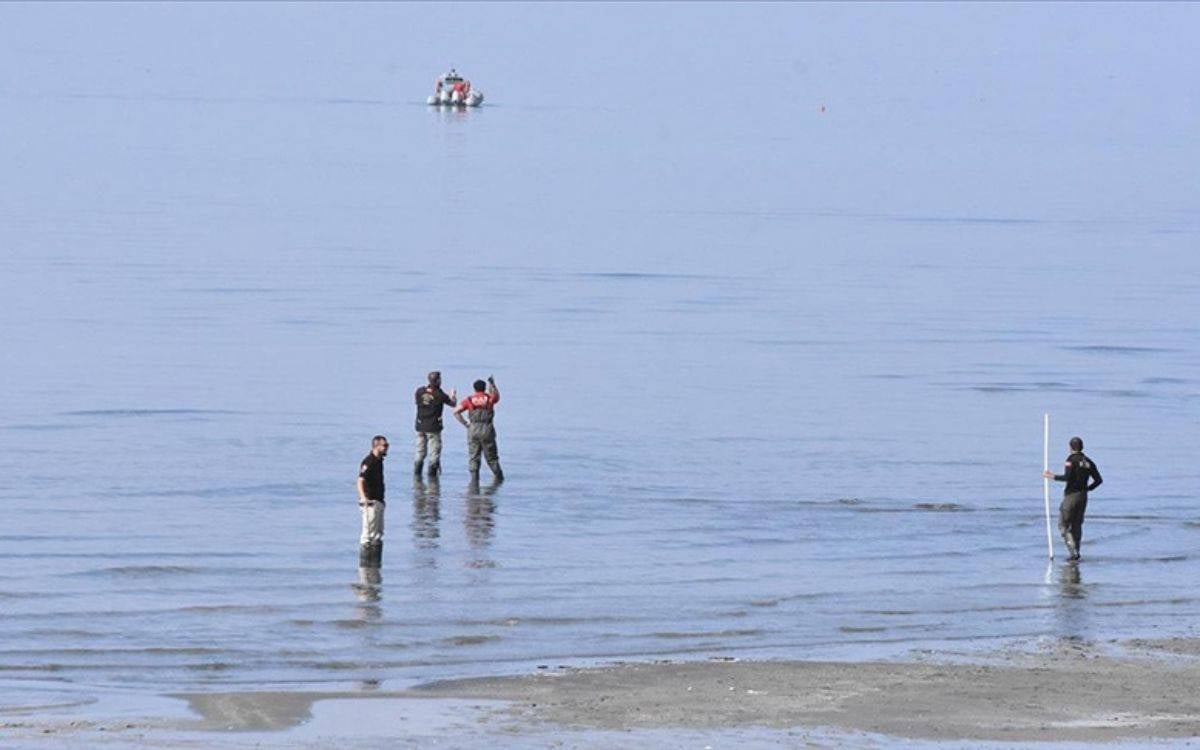 Bu fotoğraf, deniz kenarında yapılan bir arama-kurtarma çalışmasını gösteriyor. Görselde birkaç görevli, suyun içinde ve kıyıya yakın bölgede çeşitli arama faaliyetleri yürütüyor. Ekiplerden biri elinde bir ölçüm veya işaretleme çubuğu tutuyor, diğer görevliler ise su içinde arama yapıyor. Arka planda ise denizde bir bot, görevli ekip tarafından yönlendiriliyor. Aramalar, su yüzeyinde ve deniz tabanında gerçekleşiyor gibi görünüyor, deniz oldukça sakin ve geniş bir alana yayılmış durumda.