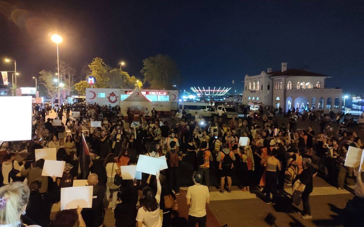 Bu fotoğraf, akşam saatlerinde kalabalık bir protesto ya da etkinlik sırasında çekilmiş gibi görünüyor. Arka planda bir meydan ya da liman alanı bulunuyor; sağ tarafta tarihi bir bina ve daha ilerde büyük bir yapı ya da stadyum aydınlatılmış şekilde görülüyor. Meydanın ortasında Kızılay’a ait bir çadır veya mobil bir stant yer alıyor. Katılımcılar ellerinde pankartlar tutarak bir araya gelmiş, birçok kişinin ellerinde beyaz pankartlar veya dövizler var. Kalabalığın arasında fotoğraf çeken ve izleyici konumunda olan insanlar da mevcut.  Mekanın bir metro durağına yakın olduğu anlaşılıyor çünkü arka planda mavi bir metro işareti dikkat çekiyor. Etkinlik, yüksek katılımlı bir protesto ya da anma etkinliğini andırıyor, gece olması nedeniyle meydanın ışıklandırması altında gerçekleştiriliyor.