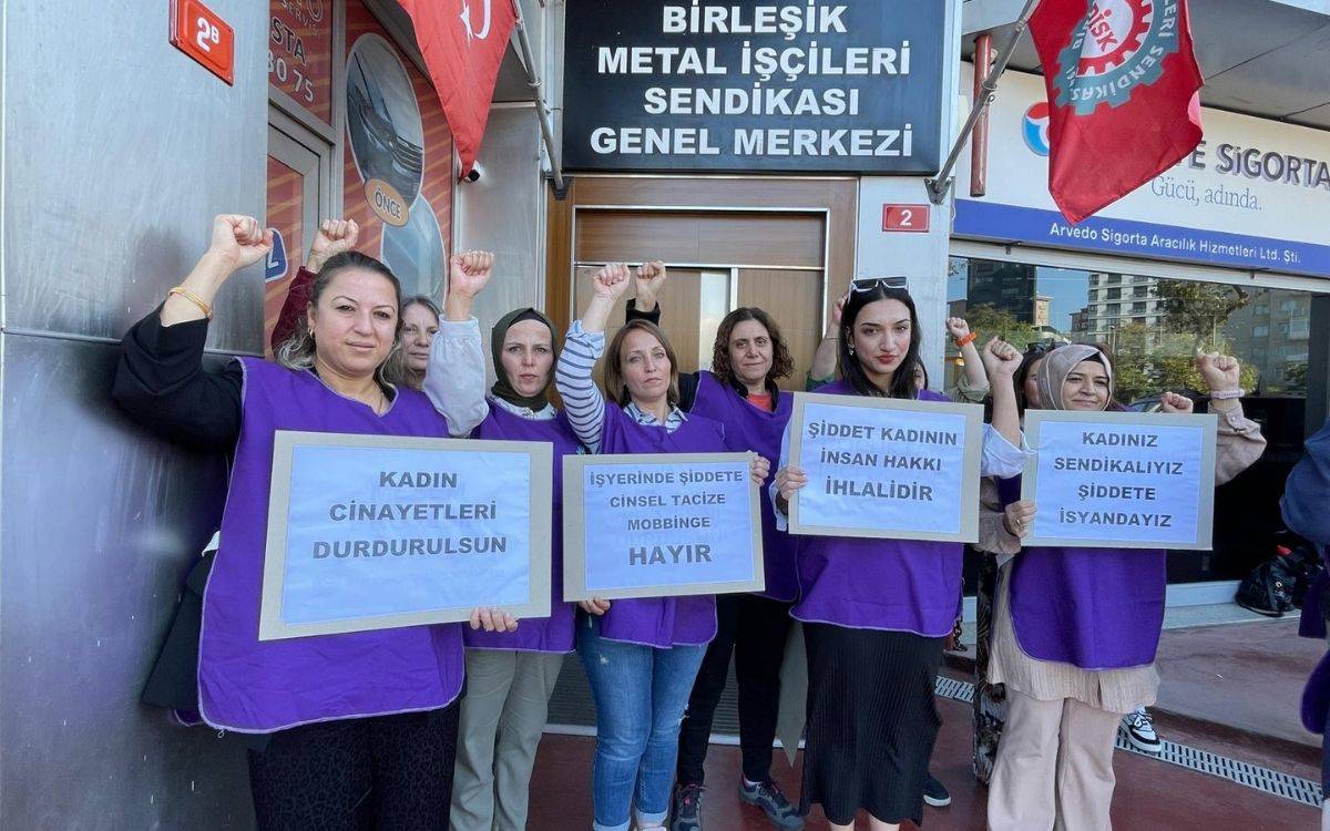 Fotoğrafta, Birleşik Metal İşçileri Sendikası Genel Merkezi'nin önünde toplanmış bir grup kadın görülüyor. Kadınlar, mor yelekler giyerek ellerinde pankartlar taşıyorlar ve birçoğu yumruklarını havaya kaldırarak protesto ettiklerini gösteriyor.