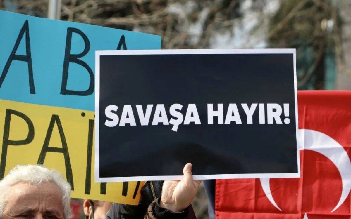 Bu fotoğrafta, bir protesto ya da miting sırasında "Savaşa Hayır!" yazılı bir pankart tutan bir kişi görülüyor. Pankart siyah zemin üzerine büyük beyaz harflerle yazılmış. Pankartı tutan kişinin sadece eli görünüyor. Pankartın sağ tarafında kısmen görünen Türk bayrağı, bu protestonun Türkiye'de yapıldığını gösteriyor.  Fotoğrafın sol tarafında ise başka bir pankartın bir kısmı daha görülebiliyor; mavi ve sarı tonlarında bir arka plan üzerine yazılmış bazı harfler dikkat çekiyor ancak tamamı okunamıyor. Önde beyaz saçlı bir kişinin bir kısmı görünmekte.  Bu görüntü, savaşa karşı yapılan bir protestoyu yansıtıyor ve barış talebini açık bir şekilde ifade ediyor. Fotoğraf, savaşa karşı çıkışın toplumsal bir mesaj taşıdığı bir anı temsil ediyor.