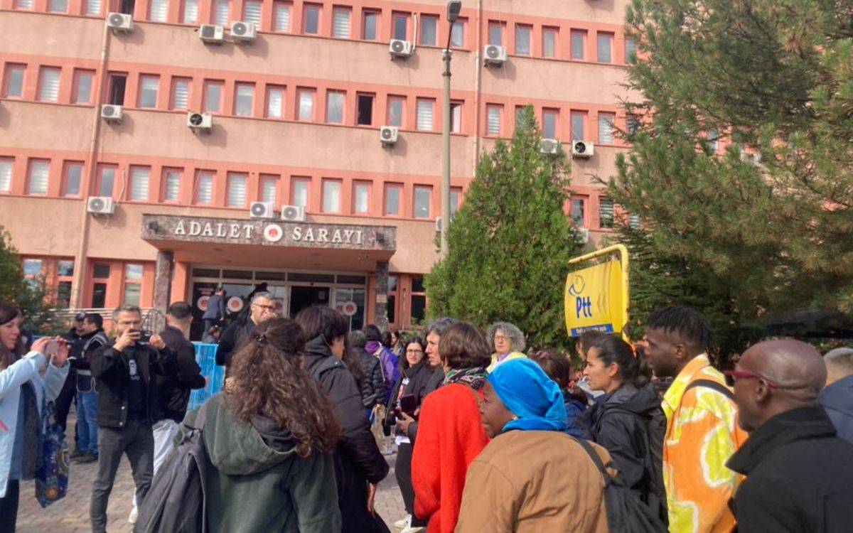 Bu fotoğrafta bir Adalet Sarayı binası önünde toplanmış bir grup insan görülüyor. Bina, birçok küçük pencereli katlara sahip ve her pencerenin altında klima üniteleri bulunuyor. Giriş kapısının üzerindeki "Adalet Sarayı" tabelası belirgin şekilde görülüyor. Ön planda, çeşitli insanlar bir araya gelmiş, bazıları telefonlarıyla fotoğraf çekiyor veya kayıt yapıyor. Gruplar halinde konuşan ve etrafı izleyen kişiler var. Arka planda bir PTT tabelası görülüyor. Katılımcılar arasında farklı etnik kökenlere ve yaş gruplarına mensup insanlar dikkat çekiyor. Kalabalık, adliye önünde toplanmış ve bir etkinlik ya da dava izliyor olabilir.