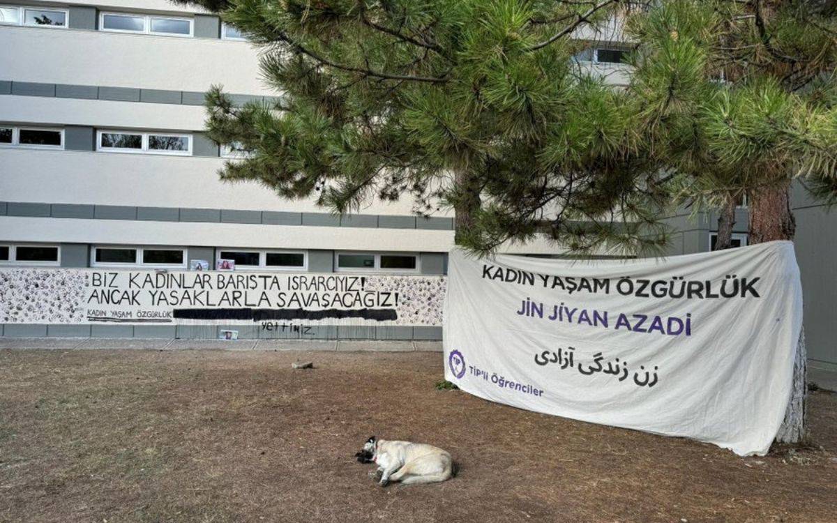 Bu fotoğraf, bir üniversite kampüsünde yapılmış bir protesto veya farkındalık eylemine ait. Görüntüde, bir bina duvarına asılmış iki büyük pankart dikkat çekiyor. Sol taraftaki pankartta, "Biz kadınlar barışta ısrarcıyız ancak yasaklarla savaşacağız!" ifadesi yer alıyor. Altında küçük puntolarla "Kadın, Yaşam, Özgürlük" yazıyor. Bu pankartın alt kısmında siyah sprey boya ile karalanmış bir kısım bulunuyor, burada "yettiniz" yazısı görünmekte.