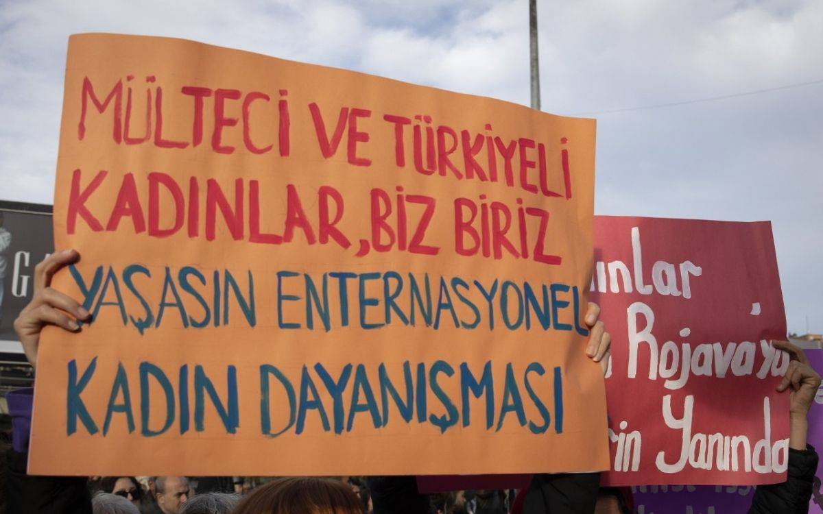 Bu fotoğrafta bir protesto veya mitingde taşınan pankartlar görülüyor. Öndeki pankartta şu ifadeler yer alıyor: "Mülteci ve Türkiyeli Kadınlar, Biz Biriz" ve alt kısımda "Yaşasın Enternasyonel Kadın Dayanışması" yazıyor. Bu pankart, kadınlar arasında dayanışma ve birlik mesajı veriyor. Arka planda ise başka bir pankartta Kürt kadınlarına yönelik bir destek mesajı yer almakta. Protesto, kadınlar arasında uluslararası dayanışmayı vurgulayan ve farklı kökenlerden kadınların bir araya gelmesini savunan bir eylemi simgeliyor.