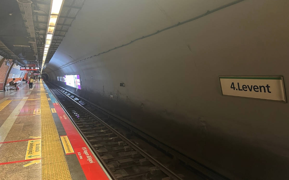 Fotoğrafta bir metro istasyonu görülüyor. İstasyonun adı, duvarda asılı bir tabelada "4. Levent" olarak yazılmış. Metro hattı boyunca uzanan raylar, ışıklandırılmış tünelin içine doğru devam ediyor. İstasyonun platformunda sarı güvenlik çizgileri ve uyarı işaretleri mevcut. Ayrıca platformun kenarına yakın bölgede kırmızı zemin üzerine yazılmış yönlendirici uyarılar ve engelli erişim alanları için işaretlemeler bulunuyor.  Solda, platformun kenarına yakın bir bankta oturan birkaç kişi var, uzakta ise birkaç yolcu metro bekliyor gibi görünüyor. Tavan kısmında aydınlatma armatürleri düzenli bir şekilde sıralanmış. İstasyonun genel atmosferi sade ve işlevsel görünüyor, şehrin bir yeraltı ulaşım sistemine ait olduğunu düşündürüyor.