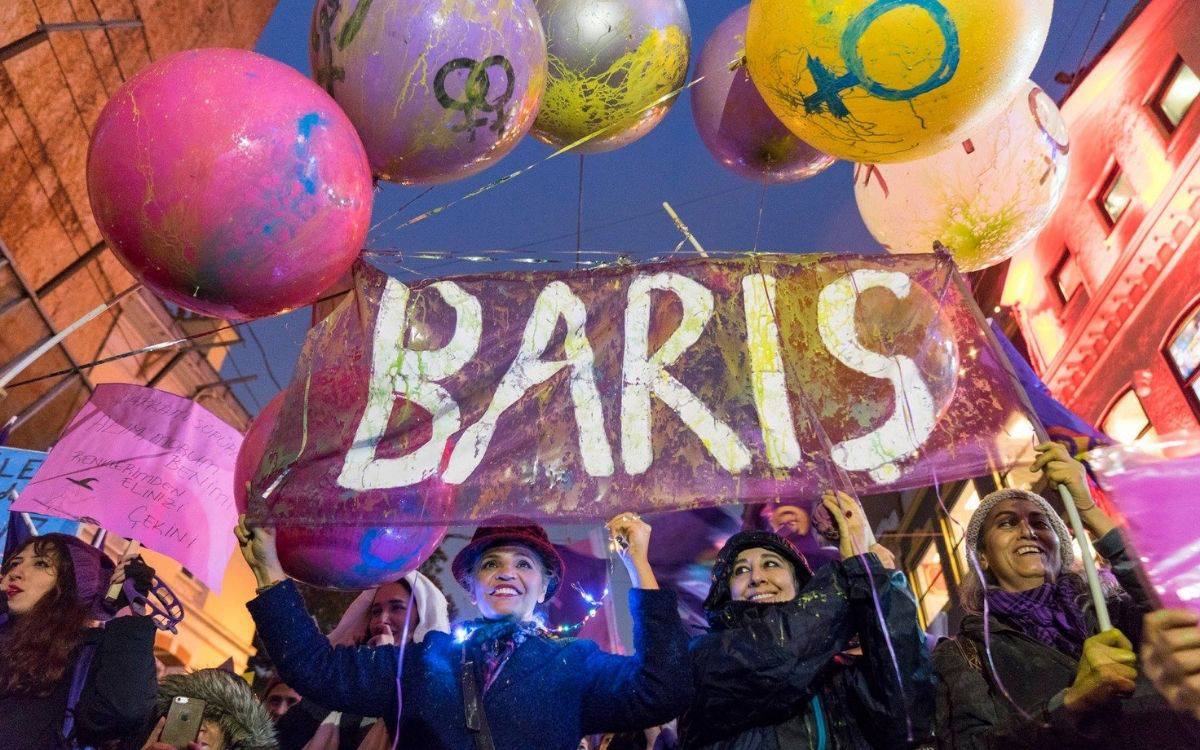 Bu fotoğraf, bir sokak protestosu ya da barış yürüyüşünden bir kareyi gösteriyor. Fotoğrafın merkezinde, üzerinde büyük harflerle "BARIŞ" yazan bir pankart taşınıyor. Pankart, parlak renklerde boyanmış ve kalabalığın elinde yükseltilmiş. Pankartın çevresinde, çeşitli renklerde balonlar var; bazı balonların üzerinde kadınlık sembolleri dikkat çekiyor. Kalabalıktaki insanlar neşeli görünüyor, çoğunun yüzlerinde gülümsemeler var. Katılımcılar rengarenk giysiler giymiş ve soğuk bir havada oldukları anlaşılıyor, çünkü bazılarının başında şapkalar ve kalın giysiler var.