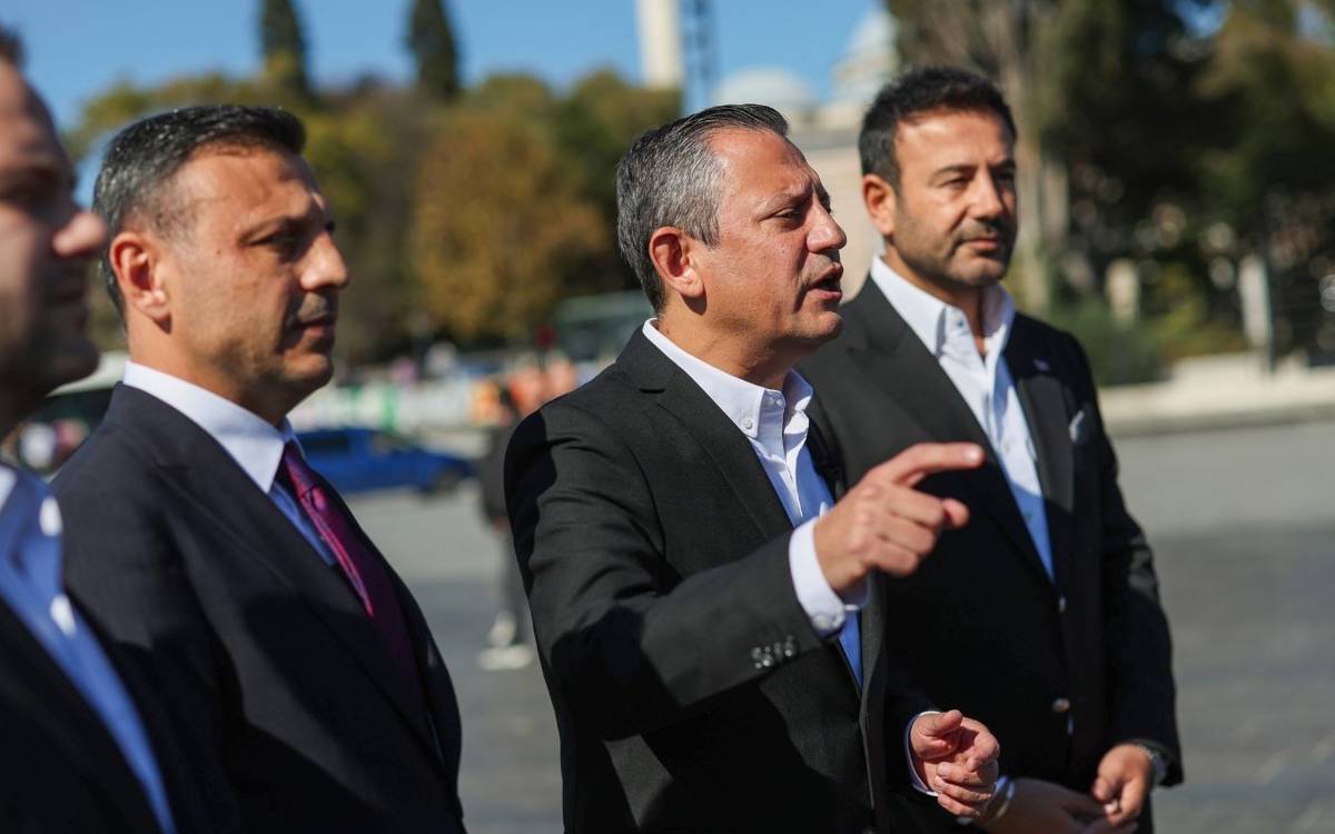 Fotoğrafta, takım elbise giymiş dört erkek açık havada bir etkinlik veya basın açıklaması yapıyor gibi görünüyor. Ön planda, yüzü ve jestleri belirgin olan kişi konuşuyor ve parmağıyla bir yeri işaret ediyor, bu da ciddi bir açıklama yaptığını veya önemli bir noktayı vurguladığını gösteriyor. Yanındaki kişiler, dikkatle onu dinliyor ve odaklanmış ifadeleriyle konuşmacının söylediklerine katılıyor gibi görünüyorlar.  Hepsi resmi v