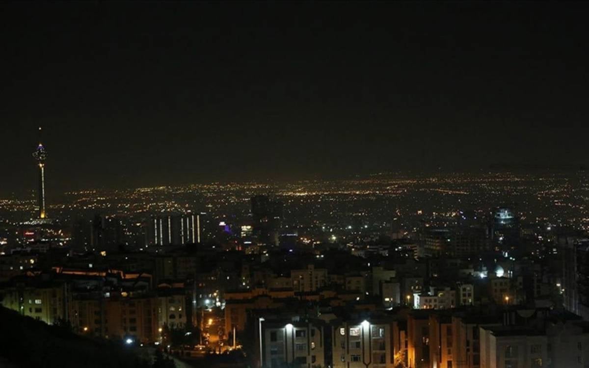 Fotoğrafta, gece vakti çekilmiş geniş bir şehir manzarası görülüyor. Şehrin ışıkları, geniş bir alana yayılmış şekilde parlıyor ve karanlık gökyüzüyle zıtlık oluşturuyor. Görüntünün sol tarafında dikkat çeken, yüksek bir kule yükseliyor; muhtemelen bu, Tahran’daki Milad Kulesi. Bu ikonik yapı şehir siluetinde öne çıkıyor. Arka planda ise şehrin ufka kadar uzanan yapıları ve ışık kümeleri göze çarpıyor. Ön plandaki daha alçak binalar ve caddelerdeki ışıklar şehre derinlik katıyor. Bu sahne, gecenin sakinliğinde Tahran’ın geniş ve yoğun yerleşim alanını betimliyor.