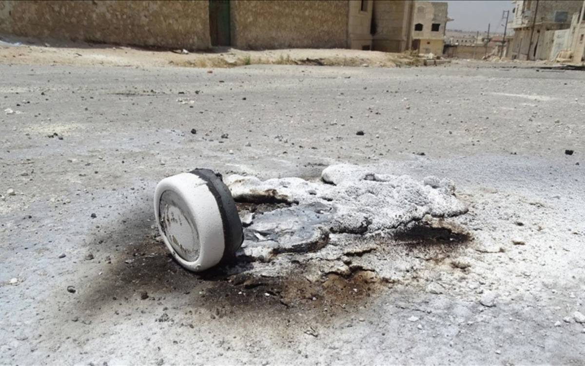 Fotoğrafta, kentsel bir alanda, bir patlama ya da yangın sonrasında oluşmuş bir kalıntı görülüyor. Yerde yanmış ve erimiş beyaz bir cisim bulunuyor. Cisim, büyük olasılıkla fosfor bombası ya da benzeri bir mühimmatın parçası olabilir; çevresindeki beyaz kalıntılar ve yanık izleri bu tür kimyasal veya patlayıcı madde kullanımını işaret ediyor. Zemin gri tonlarda ve asfalt bir yol üzerinde bu kalıntı duruyor. Arka planda ise yıkık veya hasar görmüş binalar ile boş bir sokak dikkat çekiyor, bu da çatışma ya da bombardıman sonrası bir manzara olabileceğini düşündürüyor. Fotoğrafın genel atmosferi, terk edilmişlik ve tahribat hissi veriyor.