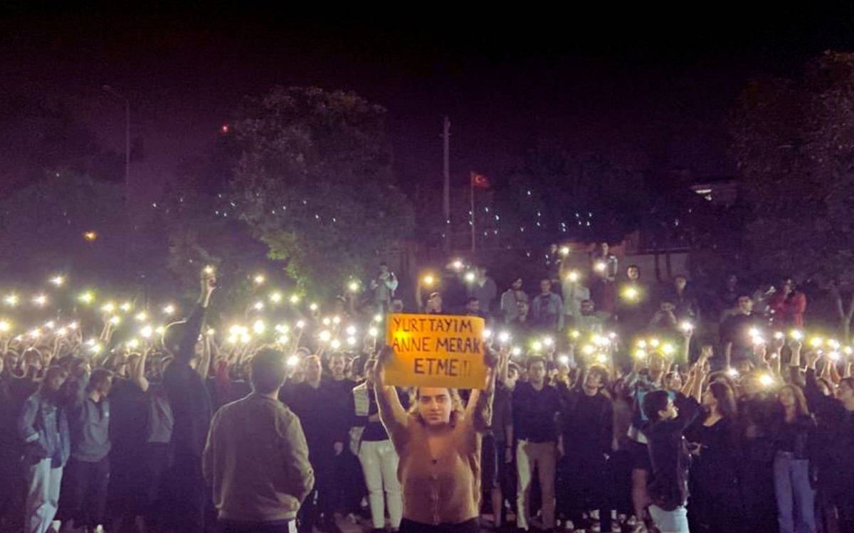 Fotoğrafta, gece saatlerinde açık alanda bir araya gelmiş büyük bir kalabalık görülüyor. Grup, telefonlarının flaş ışıklarını açarak toplu bir eylem gerçekleştiriyor. Ön planda bir kişi, turuncu bir kartona yazılmış olan "Yurttayım anne merak etme!" yazısını yukarı kaldırarak dikkat çekiyor. Bu pankart, eyleme katılan kişinin ailesine güven verici bir mesaj iletme amacında olduğunu gösteriyor. Arka planda ise Türkiye bayrağı dalgalanıyor, bu da eylemin Türkiye’de gerçekleştiğini ima ediyor. Fotoğraf, gençlerin toplumsal dayanışma ve birlik duygusunu güçlü bir şekilde yansıtıyor.