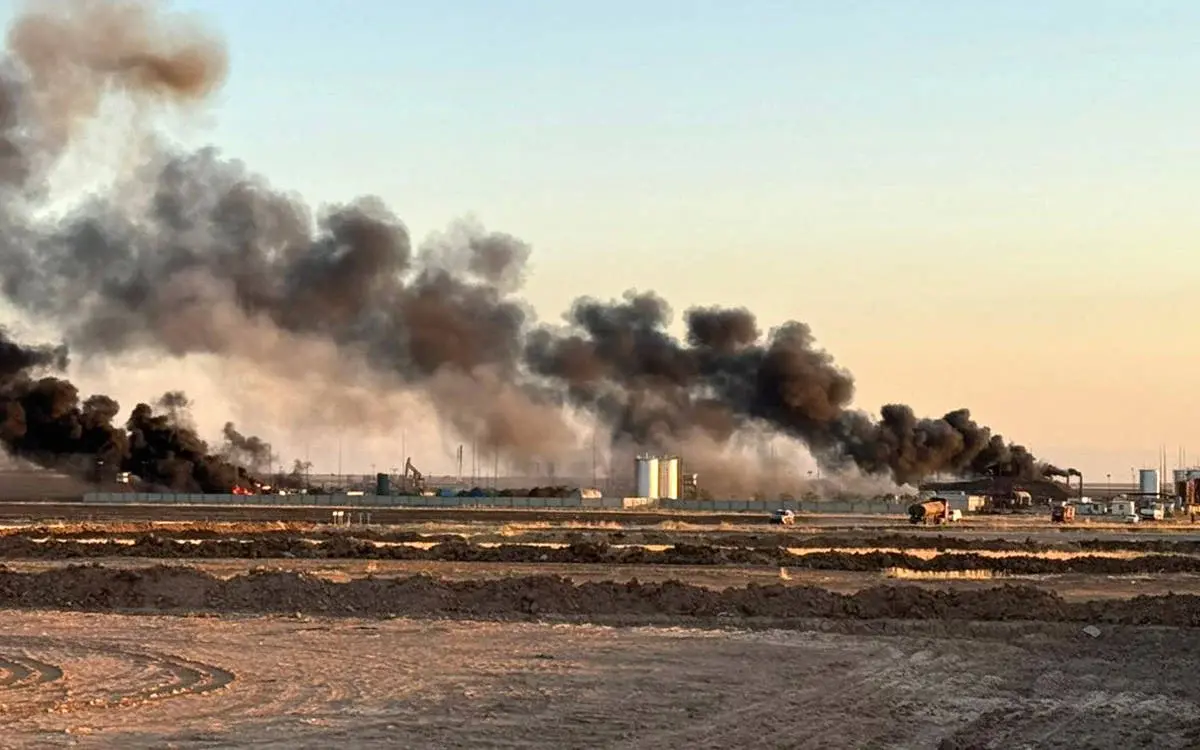 Fotoğrafta, geniş bir sanayi veya petrol sahasında yangın olduğu görülüyor. Görselde büyük bir siyah duman bulutu gökyüzüne yükseliyor. Yangının çıktığı bölge, endüstriyel yapıların ve depolama tanklarının bulunduğu bir alan gibi görünüyor. Zeminde toprak yığınları ve birkaç araç bulunuyor. Yangının etkisiyle duman oldukça yoğun ve geniş bir alana yayılmış durumda, alevler ise dumanın altında belirsiz şekilde parlıyor. Fotoğraf, güneşin batmakta olduğu bir vakitte çekilmiş gibi görünüyor; ufukta hafif sarımsı bir ışık bulunuyor. Bu durum, dumanın karanlık görüntüsünü daha da belirginleştiriyor.