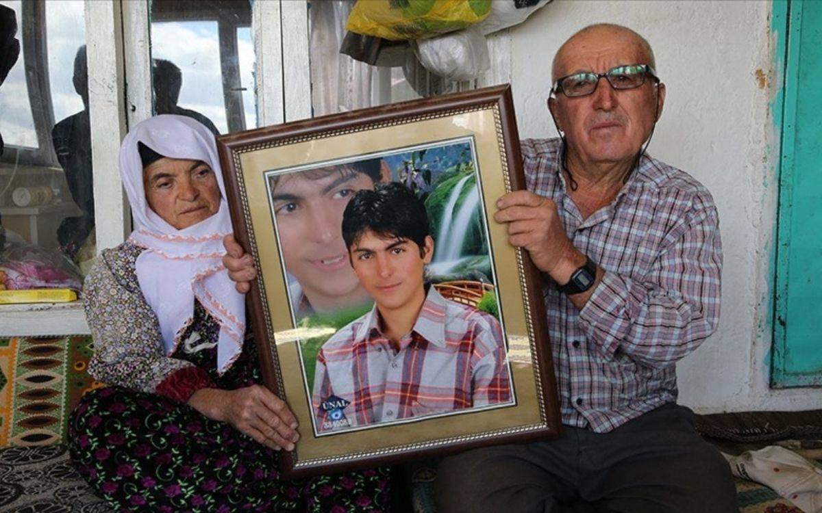 Fotoğrafta, yaşlı bir çift, kaybettikleri genç bir erkeğin büyük bir çerçeveli fotoğrafını ellerinde tutuyor. Kadın, başörtülü ve desenli bir kıyafet giymiş; gözlerinde derin bir hüzün var. Yanında oturan adam ise gözlük takıyor ve kareli bir gömlek giymiş. İkisinin de yüz ifadeleri oldukça ciddi ve üzgün, bu da büyük bir kaybın acısını hissettiklerini yansıtıyor. Fotoğraftaki genç adam, arka planda bir şelale ve doğa manzarası olan bir stüdyoda çekilmiş gibi görünüyor. Çiftin oturduğu yer sade ve mütevazı bir ortam; geleneksel motiflerle döşenmiş bir evin içi olduğu anlaşılıyor. Bu fotoğraf, kayıplarının acısını yaşayan ailelerin hüzün ve özlem dolu anısını simgeliyor.