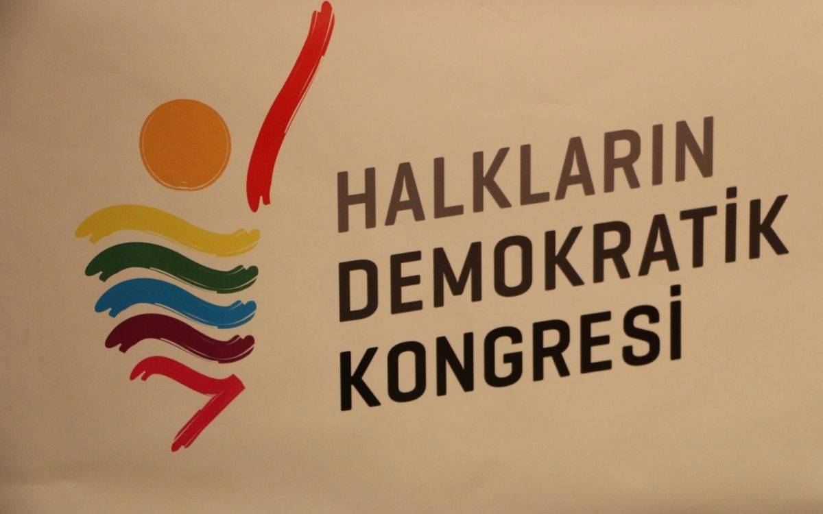 Görselde, "Halkların Demokratik Kongresi" ifadesi, büyük harflerle ve koyu siyah renkte yazılmış. Yazının sol tarafında, renkli ve soyut bir figür yer alıyor. Bu figür, kırmızı, mor, mavi, yeşil, sarı ve turuncu gibi çeşitli renklerden oluşan dalgalı çizgilerden ve yukarı doğru uzanmış bir kolu simgeleyen kırmızı bir çizgiden oluşuyor. Üst kısımda ise turuncu bir daire, güneşi veya bir başı simgeliyor gibi görünüyor. Tasarım genel olarak hareket ve birliktelik duygusunu çağrıştırıyor ve renklerin çeşitliliği, temsil edilen toplulukların farklılığını ve bir araya gelişini simgeliyor.