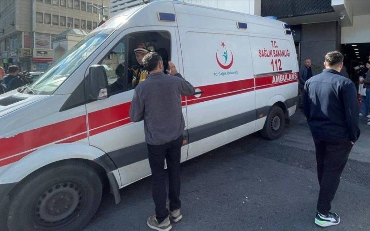 Fotoğrafta, Sağlık Bakanlığı'na ait bir 112 ambulansı görülüyor. Ambulansın ön tarafında bir sağlık görevlisi, araç içinde otururken bir kişiyle konuşuyor. Çevrede birkaç kişi daha durarak ambulansı ve olan biteni izliyor. Ambulans, şehir merkezinde bir binanın önünde park etmiş durumda ve acil bir durum veya müdahale anı olduğu izlenimi veriyor. Güneşli bir günde çekilmiş olan bu karede, ambulansın yan tarafında Sağlık Bakanlığı'nın logosu ve "AMBULANS" yazısı net bir şekilde okunabiliyor. Fotoğraf, bir acil durum müdahalesi veya tıbbi yardım anını belgelemektedir.