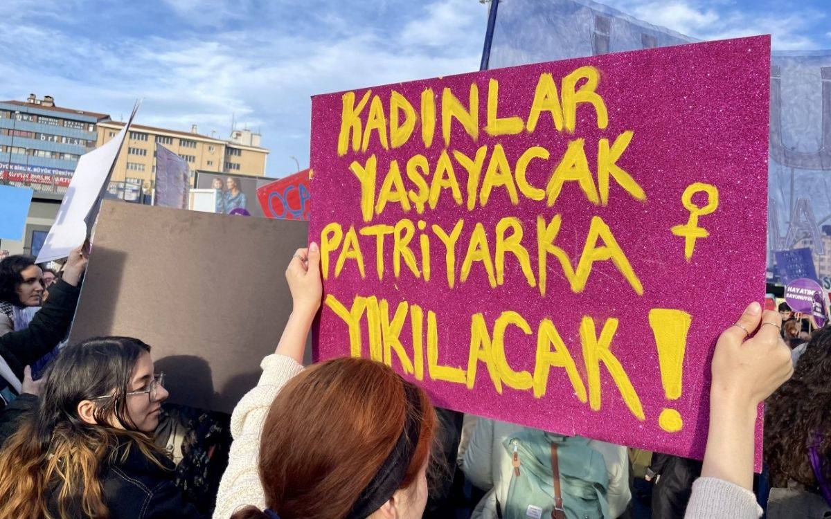 Fotoğrafta, bir protesto sırasında mor bir pankart tutan bir kadının arkadan görüntüsü yer alıyor. Pankartta büyük sarı harflerle, "KADINLAR YAŞAYACAK, PATRİYARKA YIKILACAK!" yazıyor ve kadın sembolü (♀) çizilmiş. Bu mesaj, kadınların hakları ve patriyarkal düzenin sona erdirilmesi için bir çağrı niteliğinde. Pankartın arka planında başka katılımcılar ve pankartlar görülüyor. Gökyüzü açık, hava güneşli, etkinlik açık alanda gerçekleşiyor gibi görünüyor.