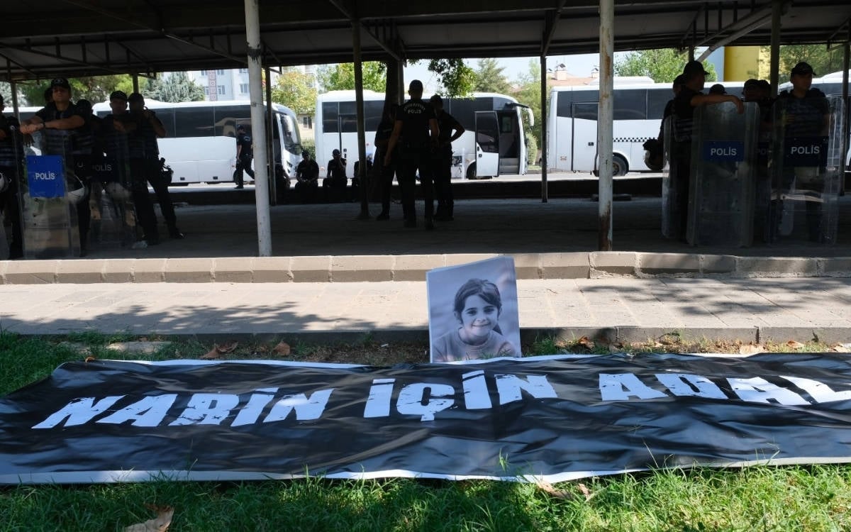 Fotoğrafta, bir park alanında polis kalkanlarıyla güvenlik önlemi almış polis memurları görülüyor. Parkın zemininde siyah bir pankart üzerinde büyük beyaz harflerle "Narin İçin Adalet" yazısı dikkat çekiyor. Pankartın hemen arkasında, genç yaşta hayatını kaybeden Narin Güran’ın siyah-beyaz bir portre fotoğrafı yer alıyor. Polis memurlarının oluşturduğu bariyerin ardında park edilmiş beyaz otobüsler var. Fotoğraf, bir protesto ya da anma etkinliği sırasında çekilmiş gibi görünüyor; polis varlığı ve pankart, olayın kamuoyunda duyarlılık uyandırdığına işaret ediyor.