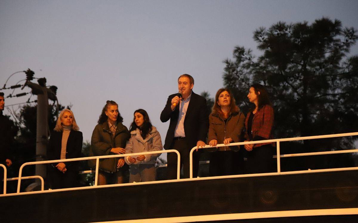 Fotoğrafta, bir grup insan bir platformda veya yüksekçe bir alanda duruyor ve konuşma yapıyor. Gecenin ilerleyen saatleri olduğu anlaşılan karede, açık hava ve loş ışıklandırma dikkat çekiyor. Grup içinde yer alan bir erkek elinde mikrofon tutarak kalabalığa hitap ediyor. Yanında çoğunluğu kadınlardan oluşan birkaç kişi daha var ve hepsi dikkatle konuşmayı dinliyor.