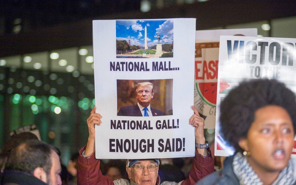 Fotoğrafta, bir protesto sırasında elinde pankart tutan bir kişi görülüyor. Pankartta üst kısımda Washington D.C.’deki ünlü National Mall'un bir fotoğrafı, alt kısmında ise Donald Trump’ın bir fotoğrafı yer alıyor. Pankartta büyük harflerle “National Mall... National Gall! Enough Said!” yazısı bulunuyor. Bu ifadeler, Trump'a yönelik eleştirel bir mesaj içeriyor ve onun cesareti veya küstahlığı eleştiriliyor.  Arka planda başka protestocular ve pankartlar da görülmekte. Kalabalık gece veya akşam saatlerinde toplanmış gibi görünüyor; çevredeki ışıklandırma bunu destekliyor. Fotoğraf, Trump yönetimine veya politikalarına karşı bir protesto etkinliğinden çekilmiş gibi duruyor.