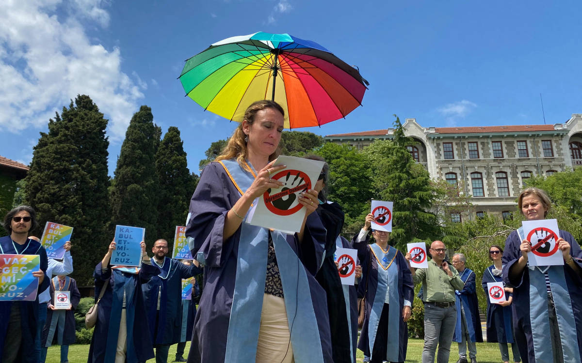 Fotoğrafta, Boğaziçi Üniversitesi'nde bir grup akademisyen, açık havada protesto ya da bir farkındalık etkinliği düzenliyor. Akademisyenlerin çoğu cübbelerini giymiş durumda ve ellerinde çeşitli pankartlar taşıyorlar. Önde duran bir kadın, renkli bir şemsiye tutuyor; bu şemsiye LGBTİ+ hareketini desteklemek için kullanılan gökkuşağı renklerine sahip. Kadın, üzerinde "Öğrencime Dokunma" yazan bir döviz tutuyor; dövizde el işareti de bulunuyor.  Arka plandaki diğer katılımcılar ise "Kabul Etmiyoruz", "Vazgeçmiyoruz" gibi ifadelerin yazılı olduğu pankartlar taşıyorlar. Etkinlik, üniversite yerleşkesinde, ağaçlar ve tarihi bir bina önünde gerçekleştiriliyor. Gökyüzü açık ve güneşli, bu da protestonun güzel bir havada yapıldığını gösteriyor. Etkinlik, akademik özgürlük, öğrenci hakları veya LGBTİ+ haklarına destek vermek gibi toplumsal bir mesaj içermekte.