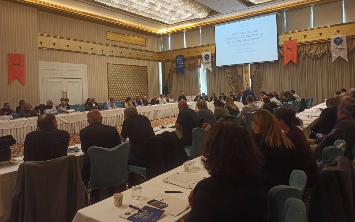 Fotoğrafta büyük bir toplantı salonunda gerçekleştirilen bir panel veya konferans oturumu görülüyor. Yuvarlak masa düzeninde toplanmış katılımcılar, konuşmacıyı dinlerken veya kendi notlarını incelerken görüntülenmiş. Arka planda projeksiyon cihazıyla yansıtılmış bir sunum başlığı yer alıyor. Sunum başlığı şu şekilde:  "Kürt Meselesinde Çözümsüzlük Türkiye'ye Neler Kaybettiriyor?"  Salonda çeşitli üniversitelerden veya kurumlardan gelen katılımcılar olduğu izlenimi veren farklı yaşlardan kişiler bulunuyor. Duvarlarda birkaç bayrak asılmış, salon klasik bir dekorasyona sahip ve gösterişli avizelerle aydınlatılmış. Katılımcılar dikkatle dinliyor veya not alıyor, etkinliğin akademik ya da düşünsel bir içerik sunduğu anlaşılıyor. Konu başlığı, Türkiye’deki Kürt meselesine dair çözüm yolları veya bu konuda yaşanan sorunların etkileri üzerine bir tartışmayı yansıtıyor.